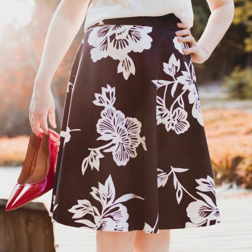 Woman holding red heels, wearing a floral skirt, enjoying a sunny day outdoors.