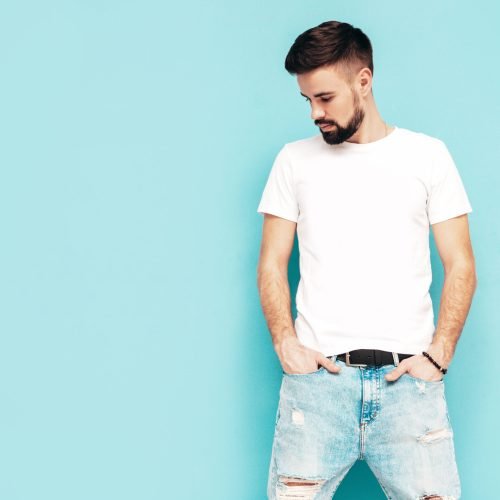 Portrait of handsome confident stylish hipster lambersexual model.Man dressed in white T-shirt and jeans. Fashion male isolated on blue wall in studio