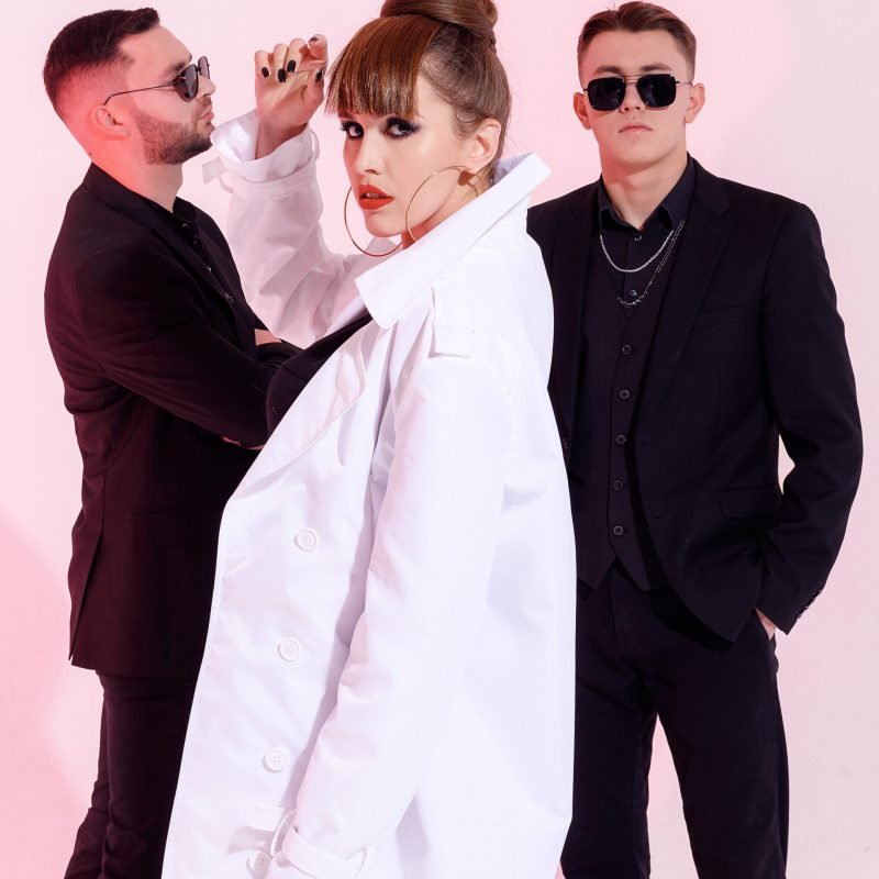 Side view of sexual female singer with beautiful makeup, wearing in white coat and jewelry, raising hand up near face and seriously looking at camera while two guys in black suits, standing behind of her on light pink background